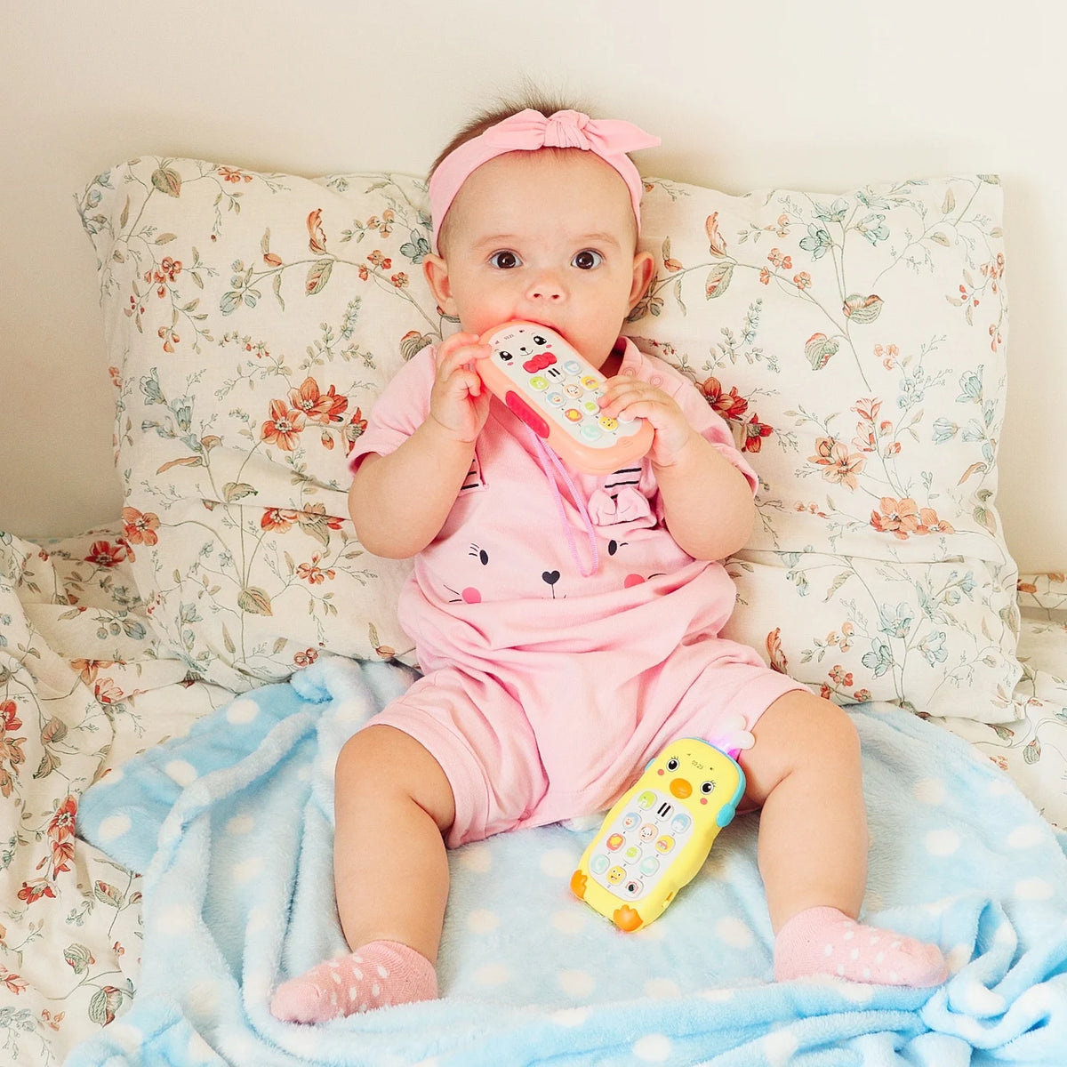 A baby wearing a pink outfit is holding a musical teether phone toy with lights and sounds.