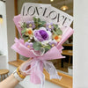 A bouquet of handmade crochet artificial flowers in purple and yellow hues wrapped in pink paper