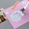 A pink felt sheet features a white layered flower cutout and a silver craft tool with an orange tag on a gray background.