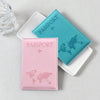 Two passport holders, one teal and one pink, featuring world maps and airplane icons, resting on a light surface.