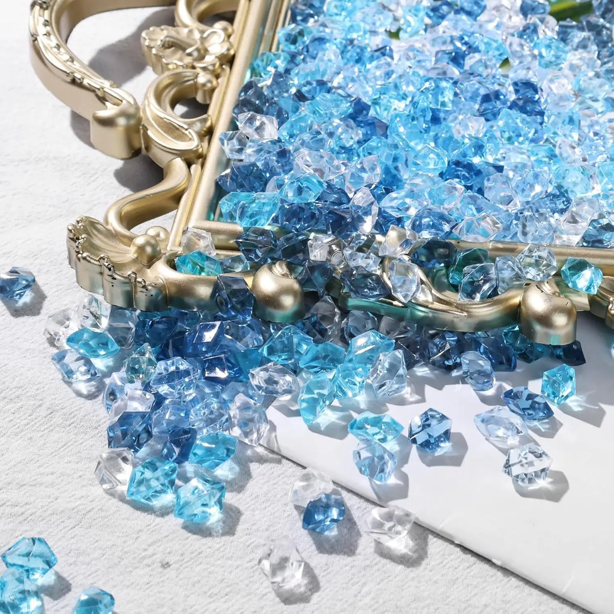Blue acrylic crushed ice gems scattered on a gold tray and white surface.