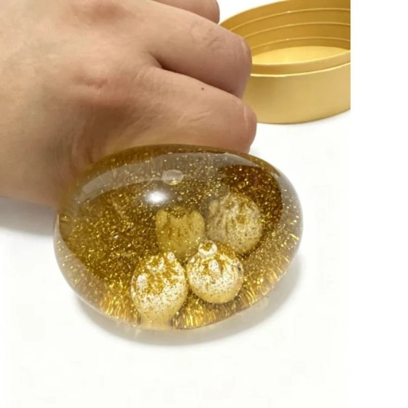 A hand holds a clear, round squishy dumpling fidget toy filled with gold glitter and small white balls.
