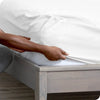 A hand is adjusting a white fitted sheet on a wooden bed frame.