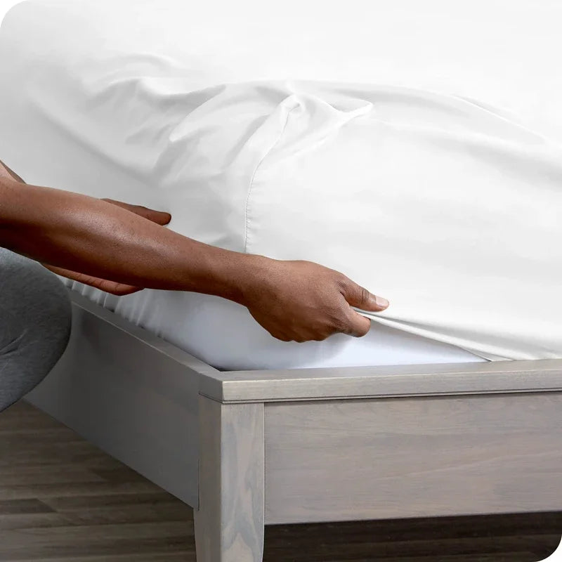 A hand is adjusting a white fitted sheet on a wooden bed frame.