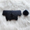 A black metal lapel pin or badge rests on a white fluffy surface next to a small black button.