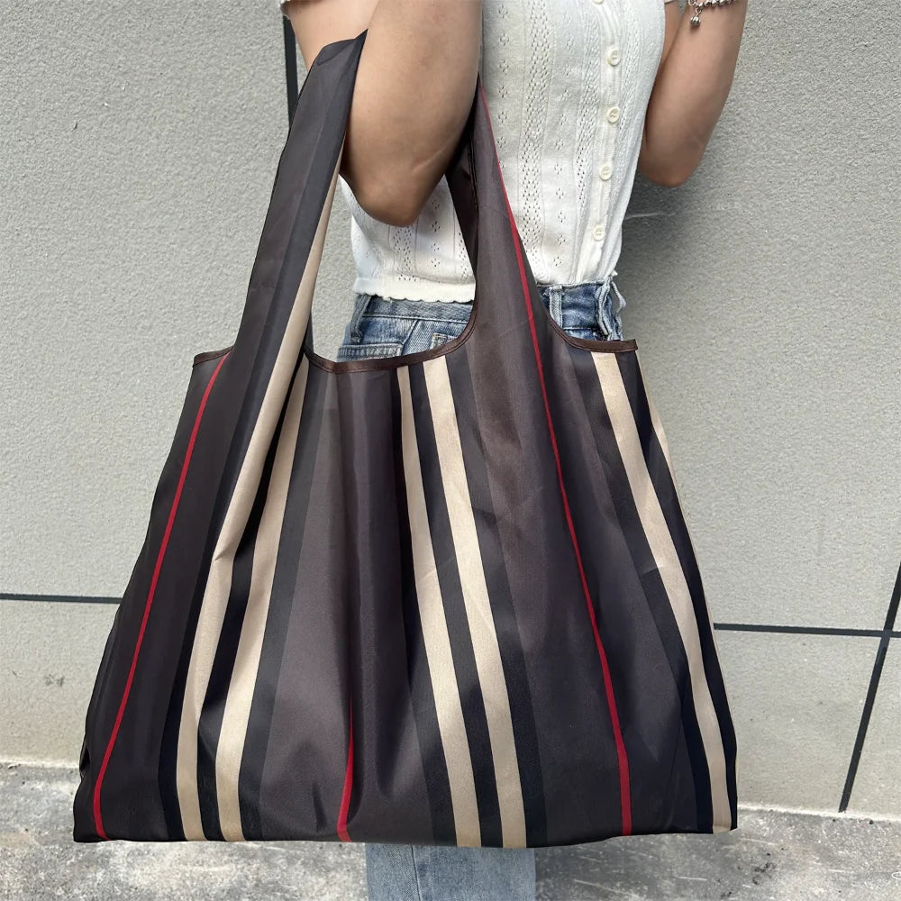 A person is holding a dark brown and beige striped foldable oxford cloth tote bag against a gray wall.