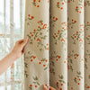 A beige blackout curtain panel with a green plant pattern is being held by a hand next to a window.