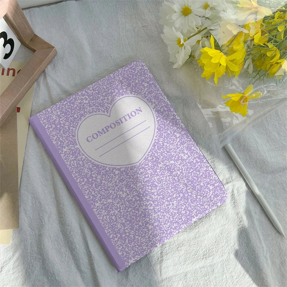 A purple composition notebook with a heart design is placed on a white cloth next to a pencil and some yellow flowers.