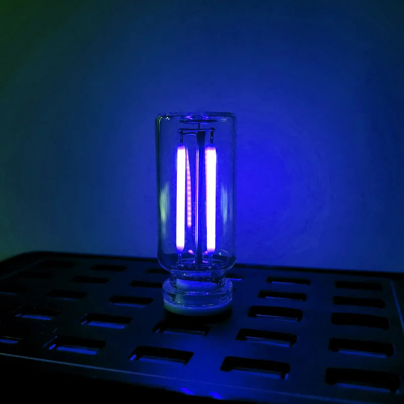 A clear glass LED night light with a warm filament glow is placed on a black tray against a blue background.
