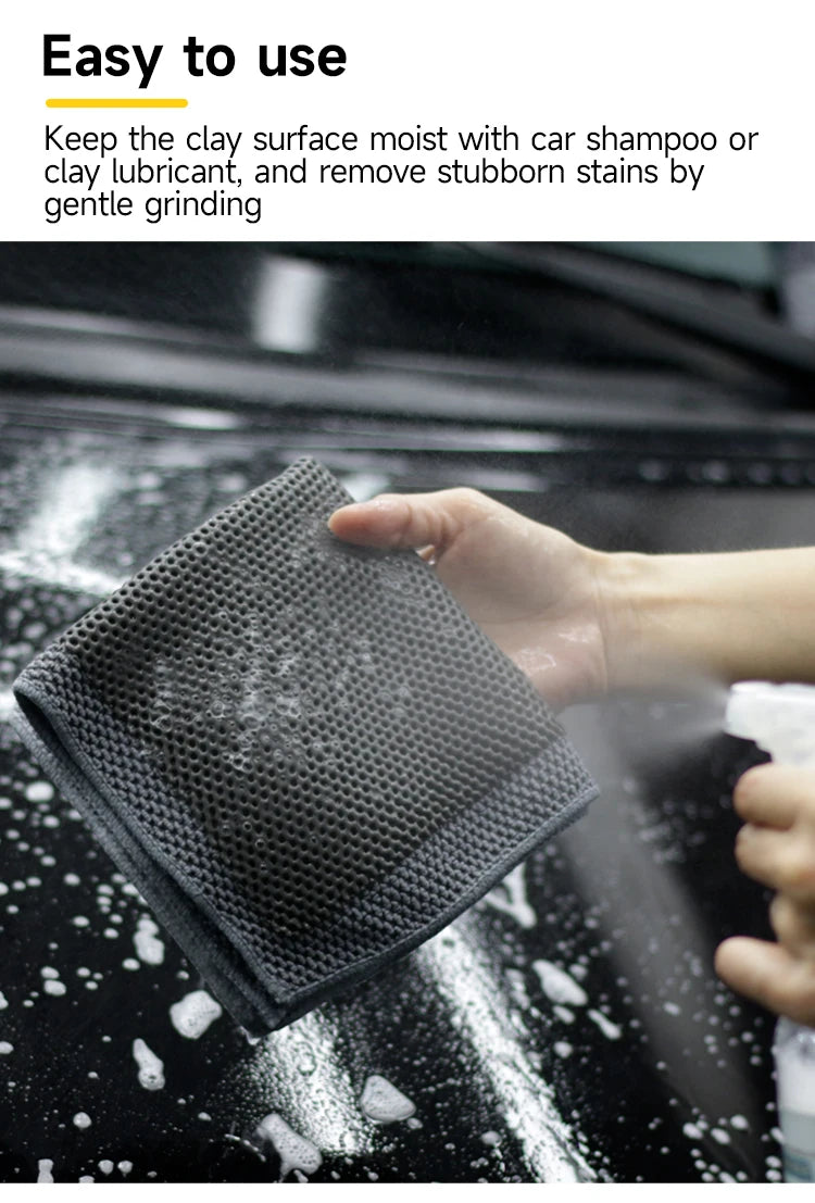 A hand is using a dark gray clay bar detailing towel to gently grind stubborn stains off a car surface.