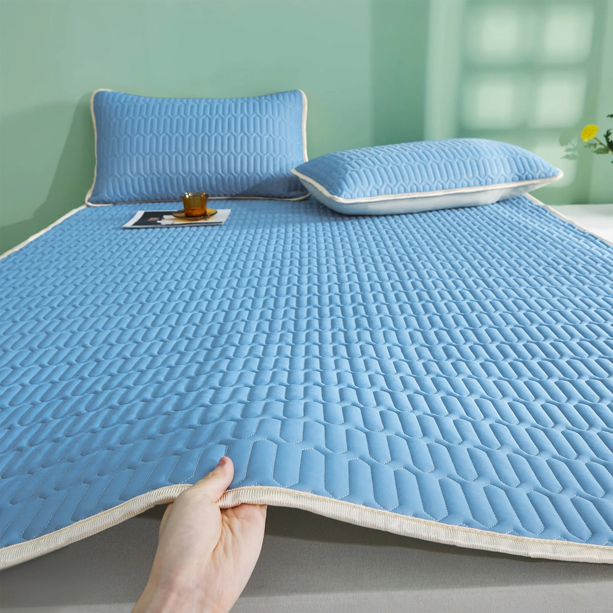 Almohadilla de colchón transpirable y refrescante azul claro mostrada en una cama con una almohada y un pequeño objeto.