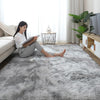 A woman sits on a gray fluffy rug reading a book next to a window with indoor plants and a TV.