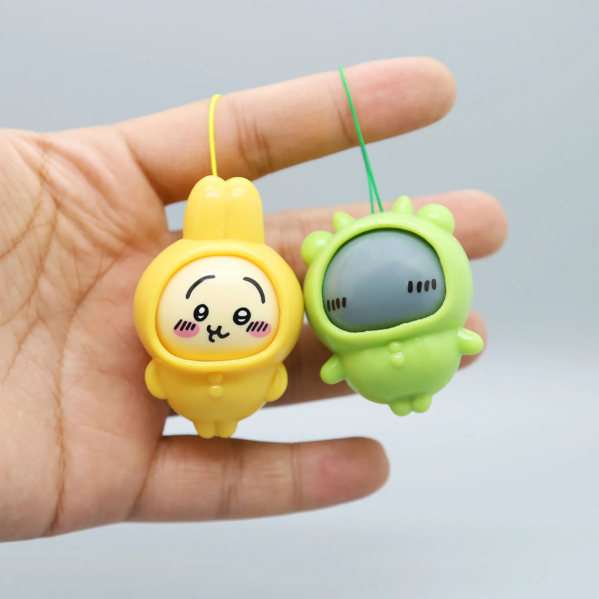 A hand holds a yellow Usagi and green Hachiware PVC bag pendant doll with string.