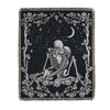 A dark fabric throw blanket featuring a white line art design of a skull, branches, and a crescent moon against a starry back