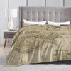 A beige plush throw blanket featuring a faded Middle Earth map design draped over a bed with a tufted headboard.