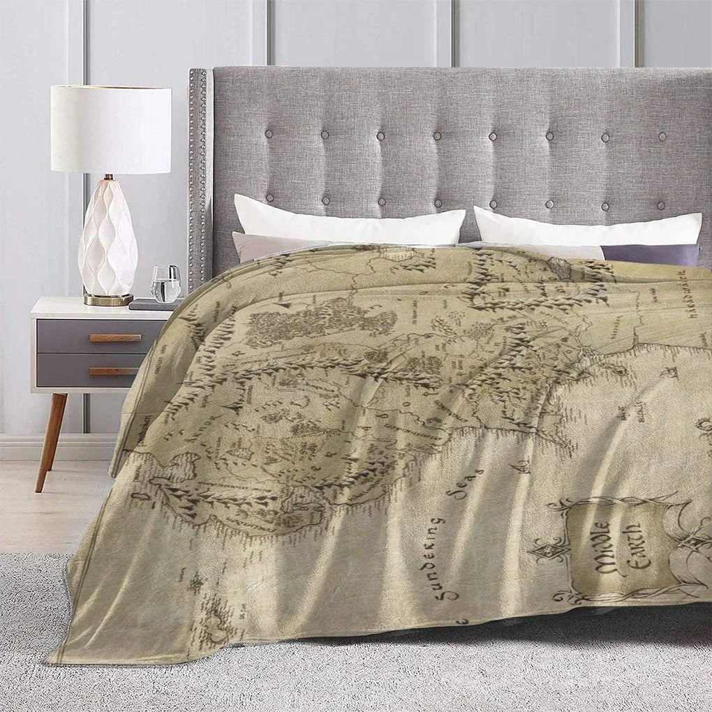 A beige plush throw blanket featuring a faded Middle Earth map design draped over a bed with a tufted headboard.