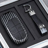 A black carbon fiber key fob cover with buttons and a silver metal key ring is displayed in a protective case.