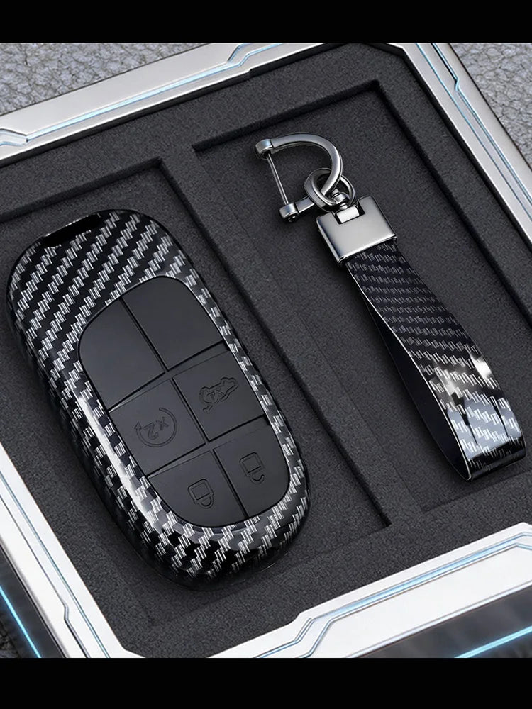 A black carbon fiber key fob cover with buttons and a silver metal key ring is displayed in a protective case.