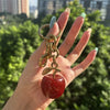 Eine Hand hält einen goldfarbenen Kristall-Obsttaschen-Anhänger-Schlüsselanhänger mit einem roten, glitzernden Apfel.