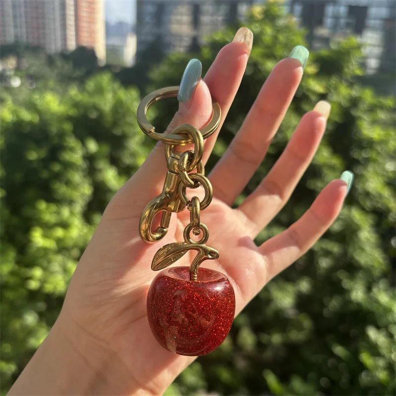 Eine Hand hält einen goldfarbenen Kristall-Obsttaschen-Anhänger-Schlüsselanhänger mit einem roten, glitzernden Apfel.