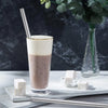 A tall glass of pinkish beverage with a stainless steel straw and cubes of white cheese on a light surface.