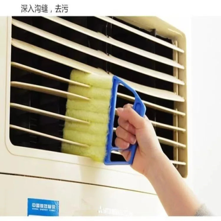 A hand is using a blue dusting brush to clean the slats of a window blind or vent.