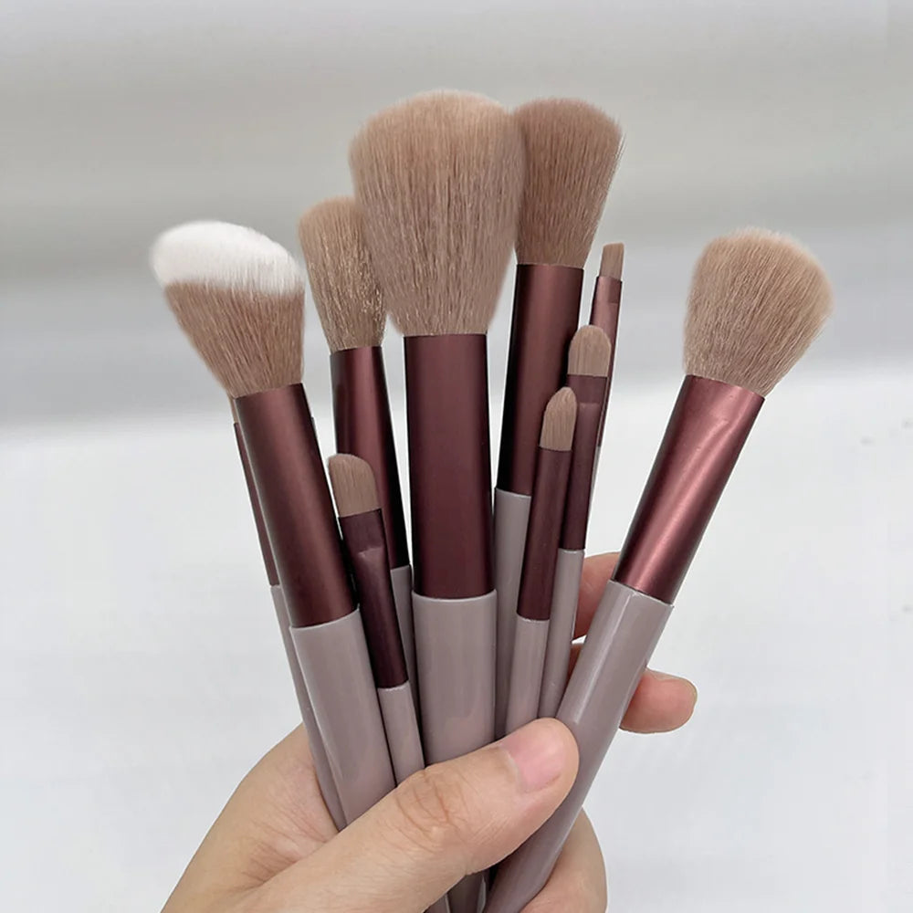 A hand holds a set of thirteen makeup brushes with natural hair and rose gold handles.