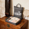 A dark gray laundry bag rests on a wooden trunk next to a tall wooden object.