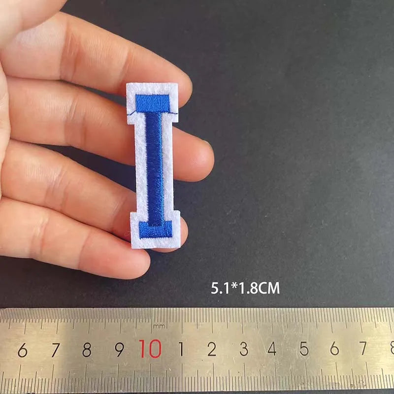 A hand holds a blue embroidered letter I patch, measuring 5.1 by 1.8 cm against a ruler.
