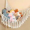 A bohemian plush toy storage hammock made of white macrame is hanging against a beige wall, holding various stuffed animals.