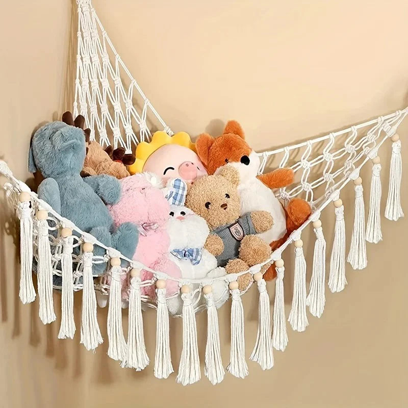 A bohemian plush toy storage hammock made of white macrame is hanging against a beige wall, holding various stuffed animals.