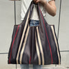 A dark brown and beige striped foldable oxford cloth tote bag is being held by a person against a light gray background.
