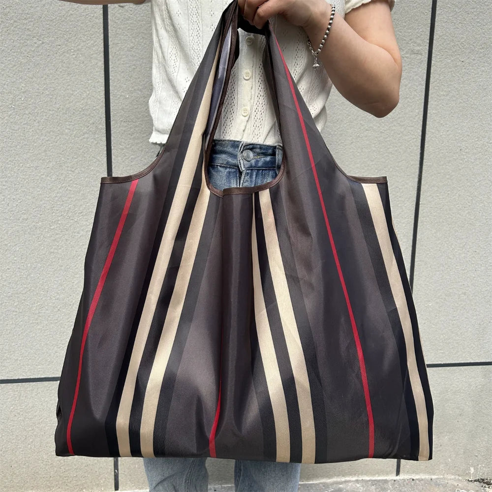 A dark brown and beige striped foldable oxford cloth tote bag is being held by a person against a light gray background.