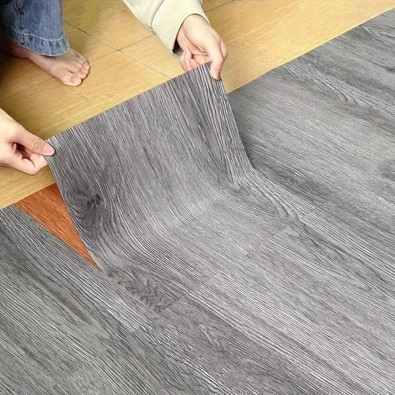Hands are peeling a sheet of gray wood grain vinyl flooring tiles onto a light wooden floor.