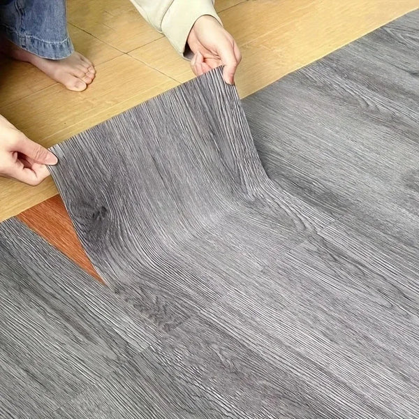 Hands are peeling a sheet of gray wood grain vinyl flooring tiles onto a light wooden floor.