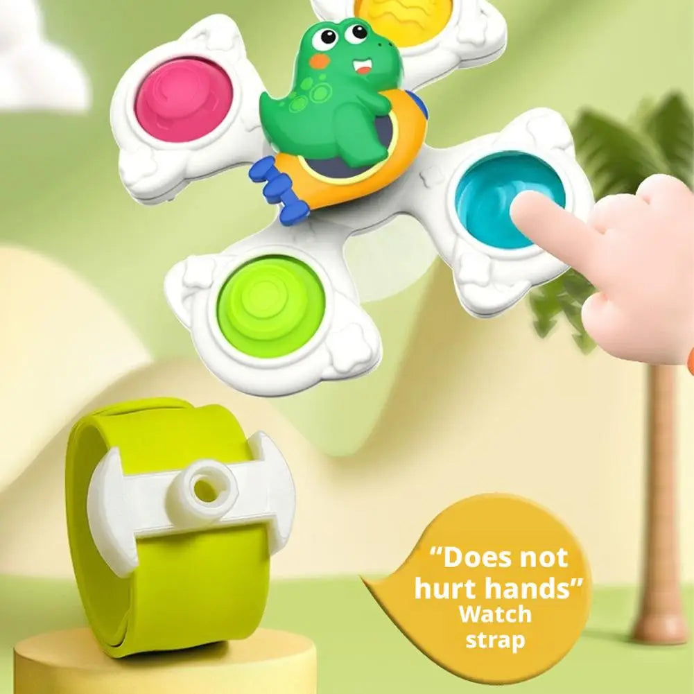 A hand is interacting with a white suction cup spinner toy featuring a green dinosaur and an orange bead.