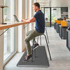 A man sits on an anti-fatigue standing mat at a modern desk working on a laptop.