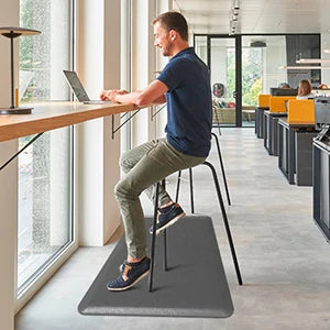 A man sits on an anti-fatigue standing mat at a modern desk working on a laptop.