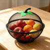 A black metal fruit basket with a green mesh cover filled with various fresh fruits like apples and oranges.