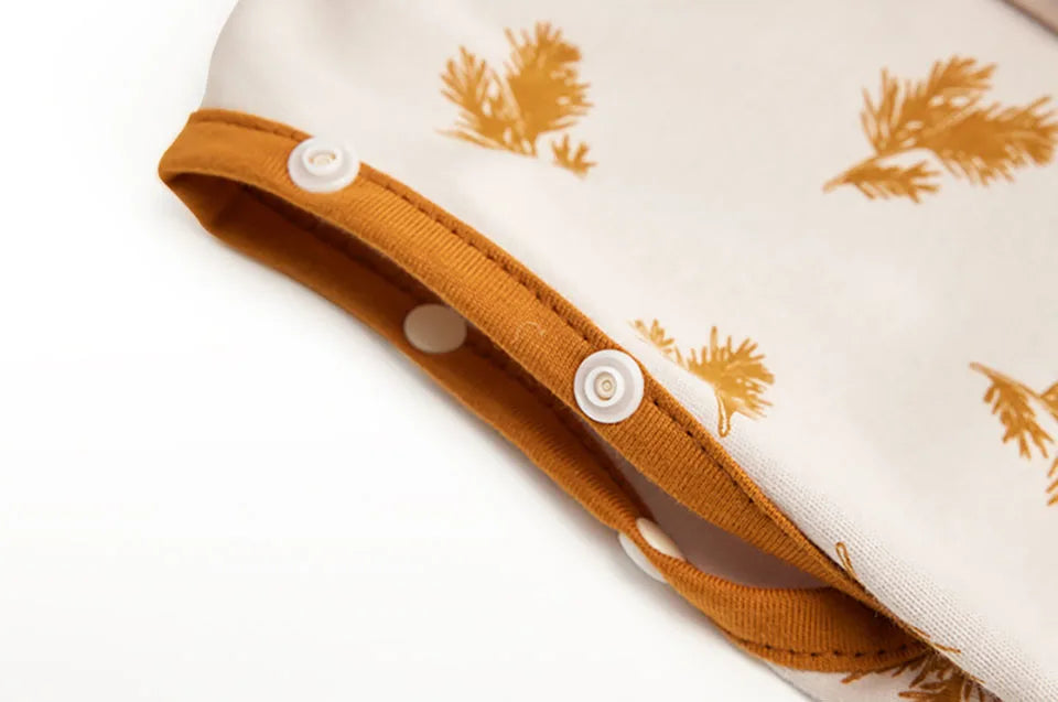 A close-up of a beige baby sleep sack with a mustard yellow trim and embroidered pine branch pattern.
