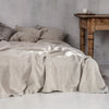 A light beige linen bed sheet set draped over a bed with pillows, next to a rustic wooden table.