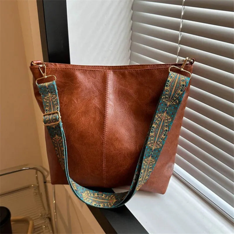 A brown leather shoulder bag with golden embroidery hanging in front of a white window blind.