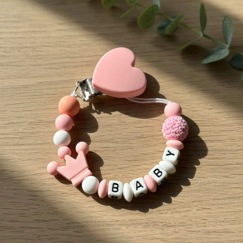 A pink and white beaded pacifier clip bracelet with a heart charm rests on a wooden surface.