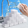 A hand is squeezing a white melamine cleaning sponge under running water, with a stack of similar sponges in the foreground.