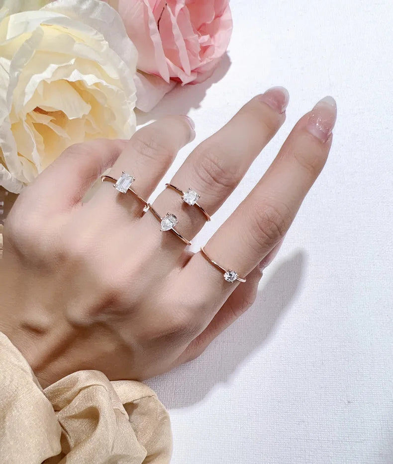 A hand wearing three minimalist moissanite solitaire rings on a white background with pink and yellow flowers.