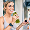 A woman holds a green juice bottle, smiling, in a bright indoor setting.