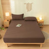 A brown fitted bed sheet set with matching pillowcases is displayed on a wooden bed frame in a bright bedroom.