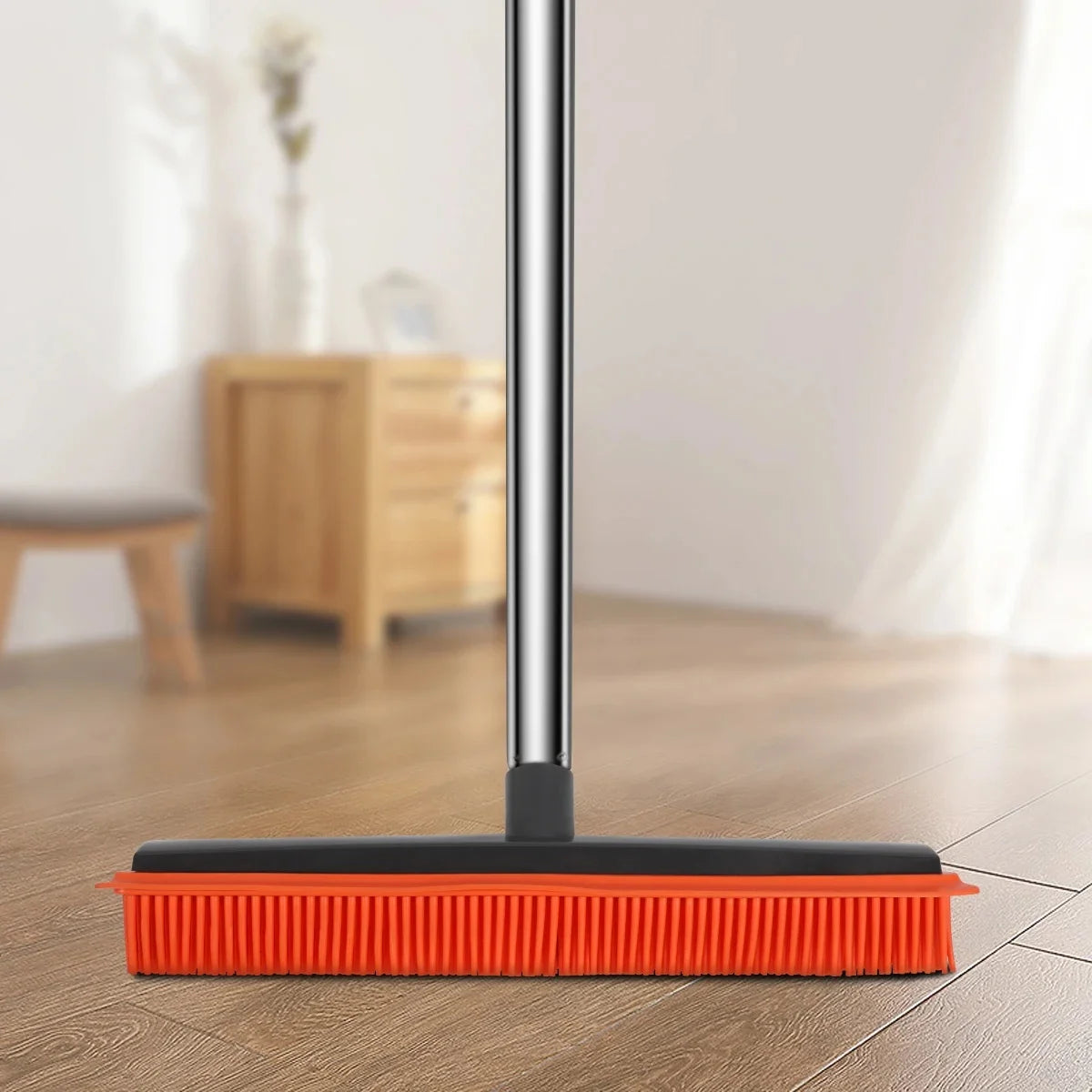 A rubber broom and carpet rake with an adjustable silver handle resting on a wooden floor.