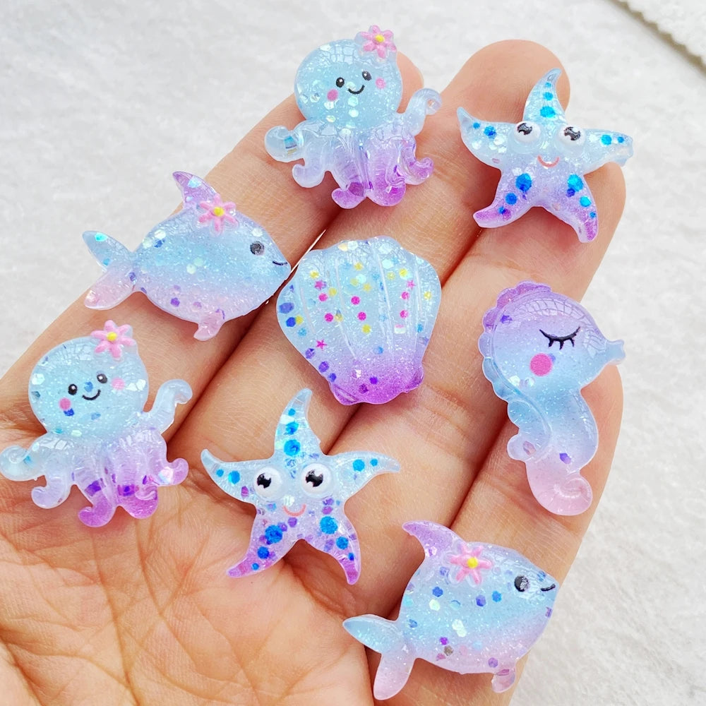 A hand holds several iridescent, glowing blue and purple resin charms shaped like sea creatures for crafting.