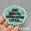 A teal embroidered patch with black text reading ONE MENTAL BREAKDOWN LATER and a gold crown detail, held in a hand.